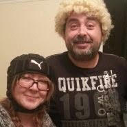 Tall light-skinned man in a printed tee shirt with blonde curly hair next to a shorter brown haired woman in glasses and a knit cap.