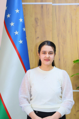Young Central Asian woman in a white blouse and black pants or skirt standing in front of a wooden wall and a flag.