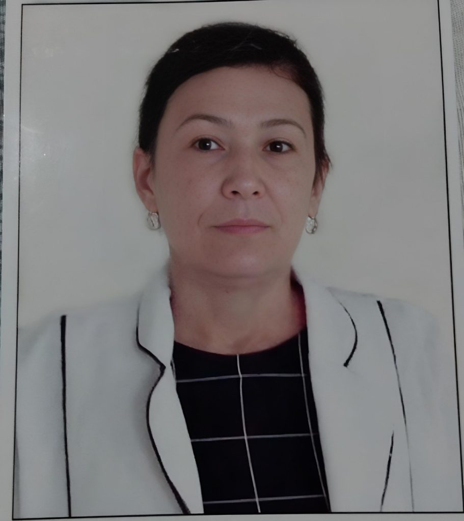 Central Asian woman with dark hair up in a bun and small earrings and a tan coat and black blouse. 