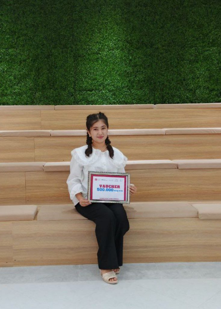 Central Asian teen girl with two dark braids, a white ruffled blouse, and an award in her lap, seated on a wooden step in front of green ivy leaves. 