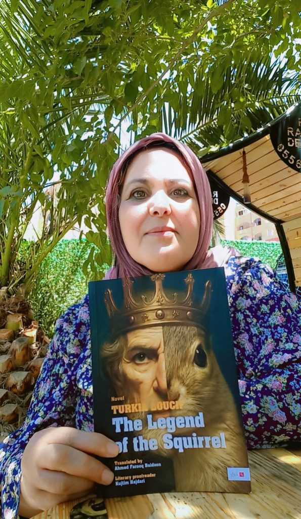 Young middle aged Arab woman with a pink headscarf and flowered blouse standing under a tree on a sunny day holding a copy of her book with the head of an old king and a squirrel side by side. 