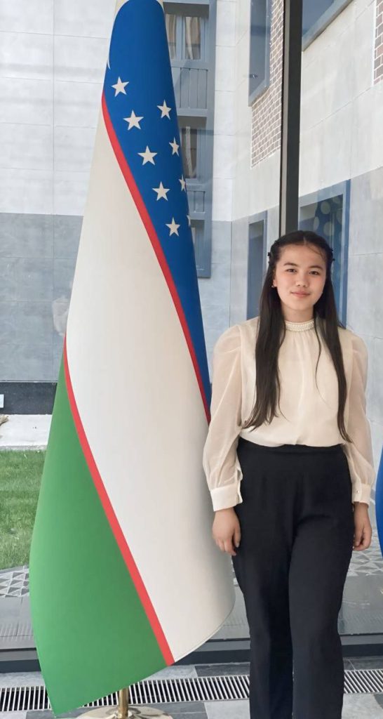 Young Central Asian woman with long dark hair in a white blouse standing next to the Uzbek flag.