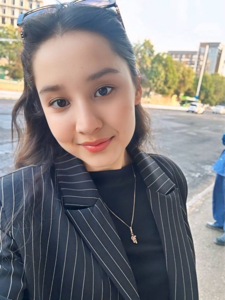 Young Central Asian woman leaning to the left. She's got long curly dark hair, brown eyes, and a striped black vest over a black blouse with a small necklace. She's on a city street with trees and a sidewalk and buildings in the distance.