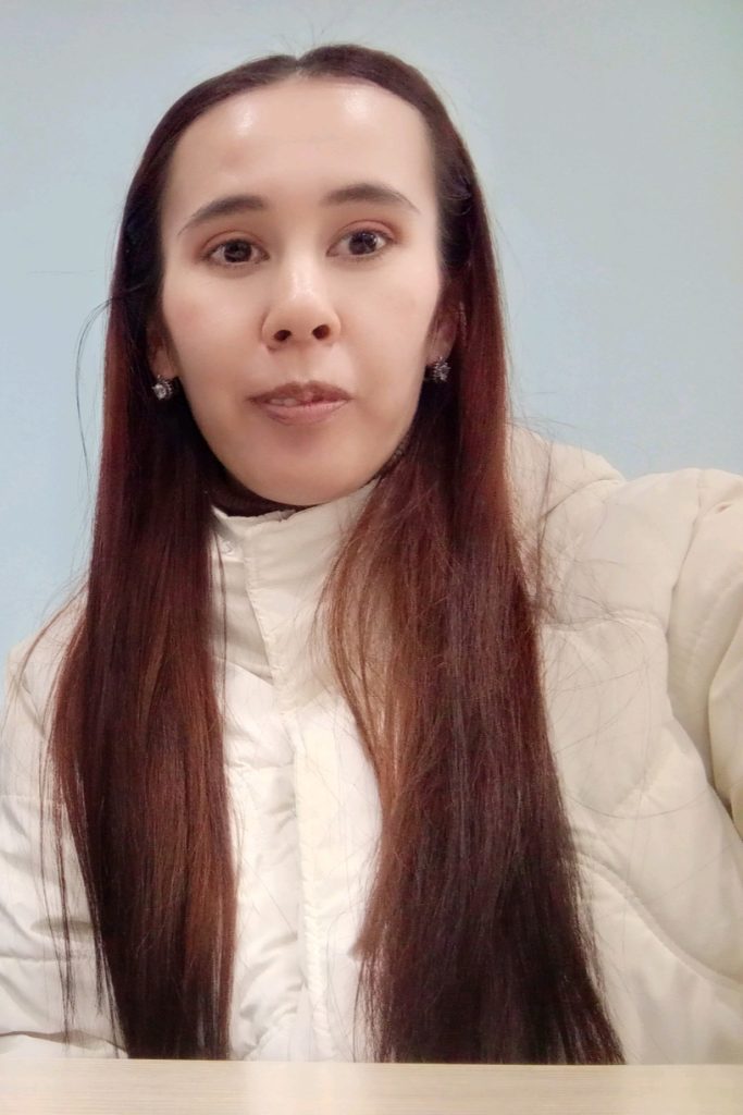 Young Central Asian woman with dark reddish-brown hair, small earrings, and a tan poofy jacket.