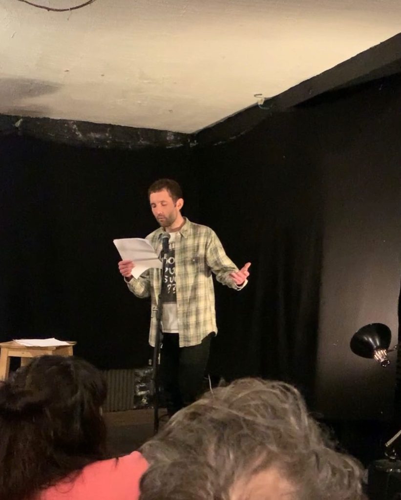 Young white man with short brown hair and a plaid jacket over a tee shirt with words on it reads poetry into a microphone on a small stage. 