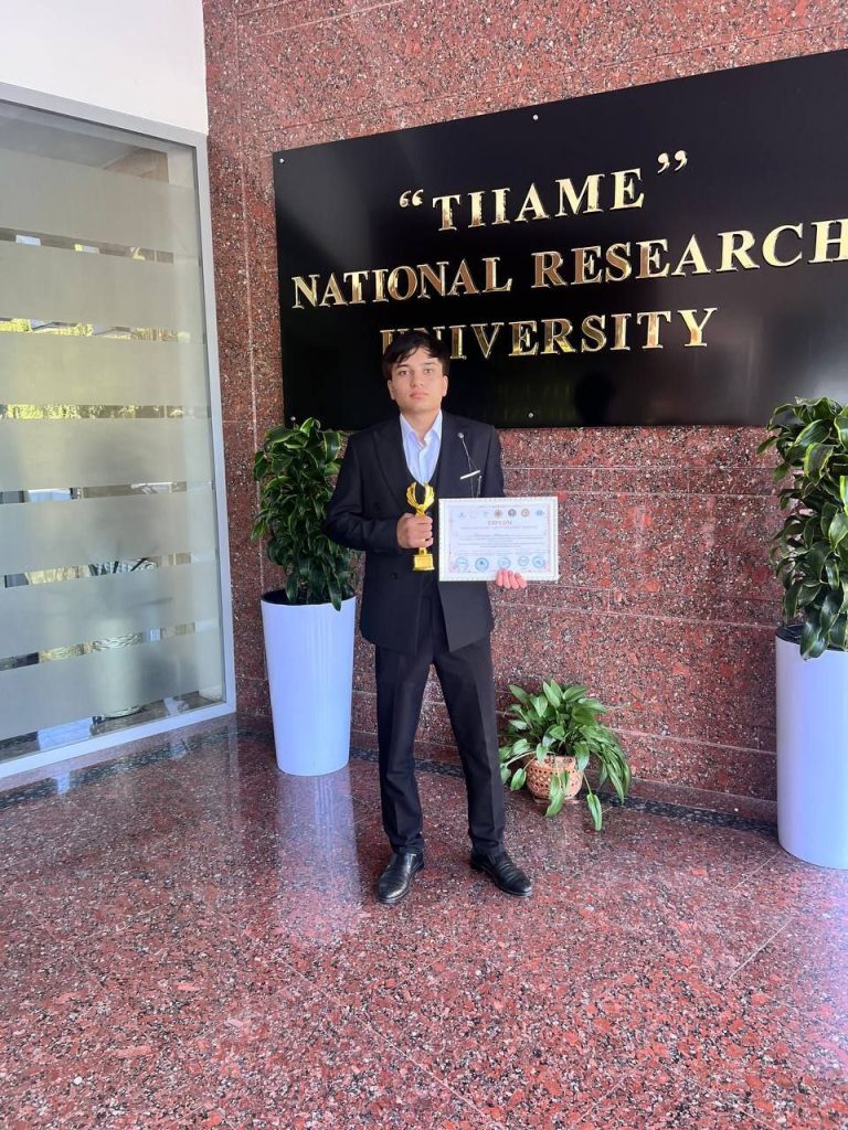 Young Central Asian man holding an award and certificate in front of a sign for his university. He's in a collared shirt and dark coat.