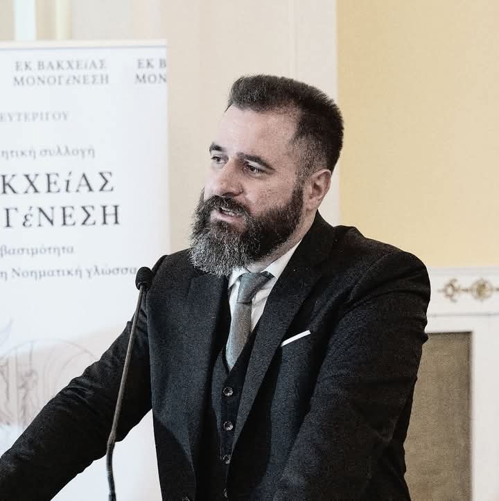Older middle aged man with a trimmed beard and hair in a black suit and tie speaking into a microphone. Greek words behind him.