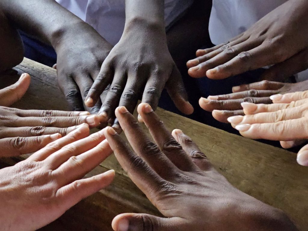 Hands of different shades of skin color meeting palms down over a wooden board. 