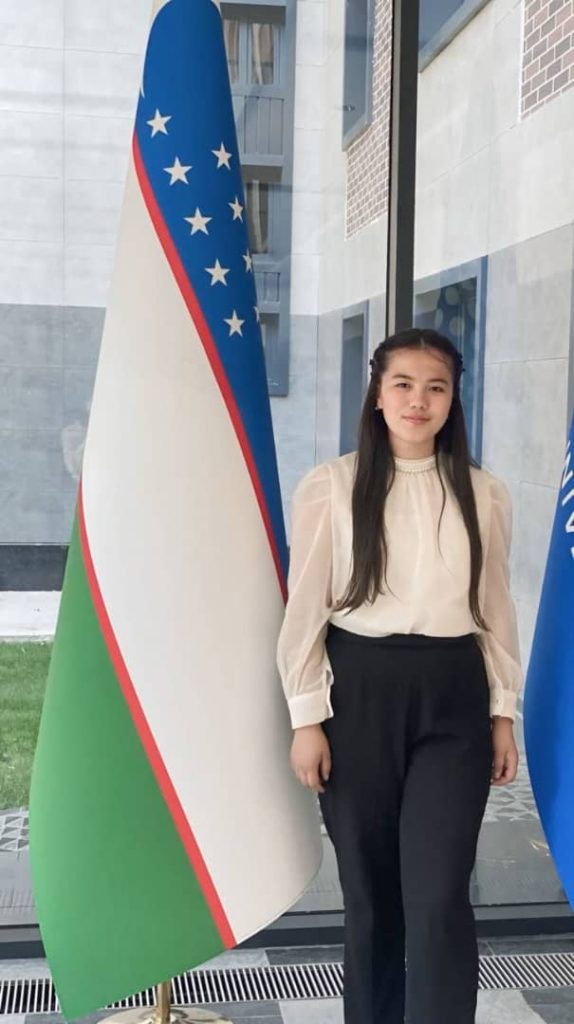 Central Asian teen girl in a white blouse and black skirt in front of the Uzbek flag. 