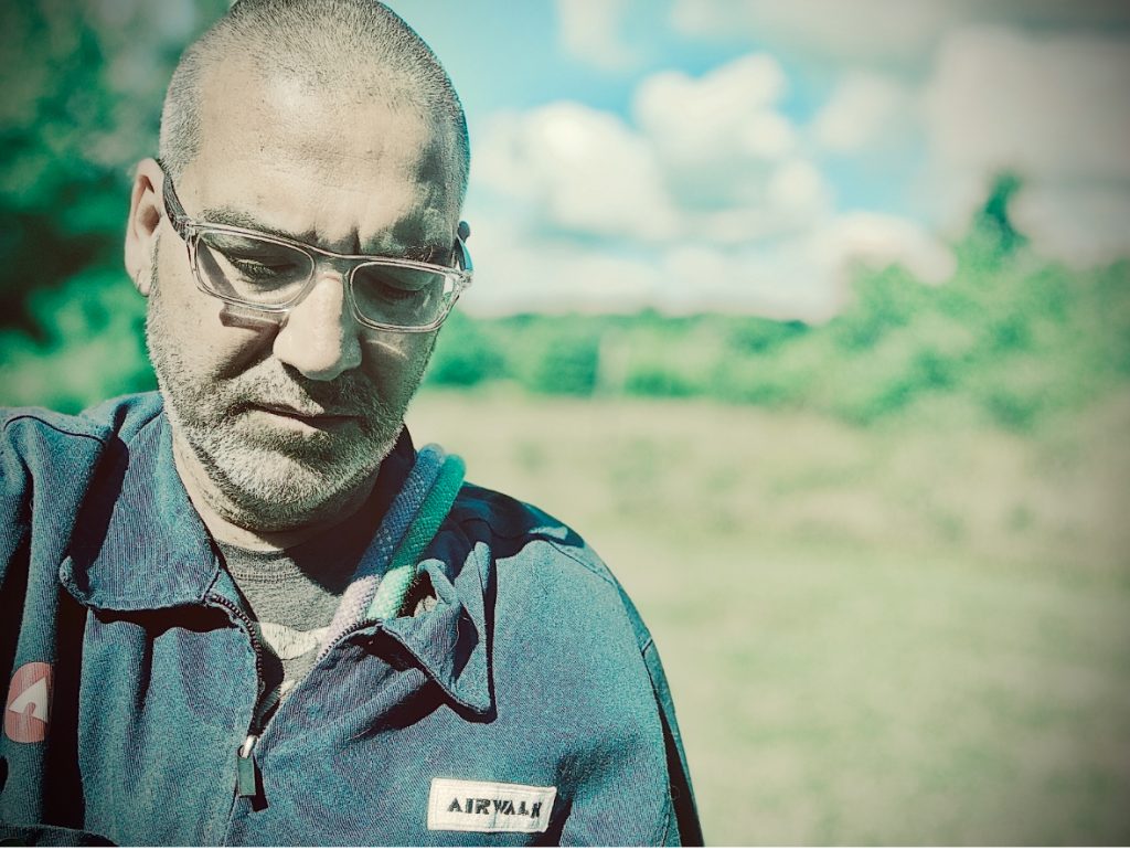 Middle aged light skinned man in reading glasses and a zipped up jacket over a tee shirt, looking down. Trees and grass and clouds in the distance.