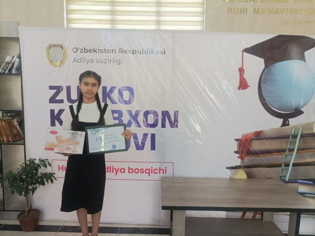 Central Asian teen girl with two braids, a black dress over a white blouse, holding certificates in front of a banner in a classroom with a globe and books and a cap. 