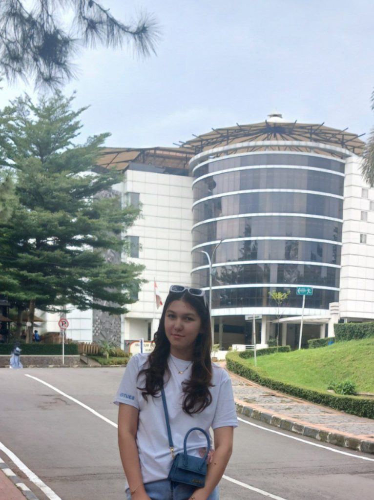 Central Asian woman with sunglasses, a tee shirt, a black purse and blue jeans in front of buildings with big semicircular windows and trees and grass and a road. 