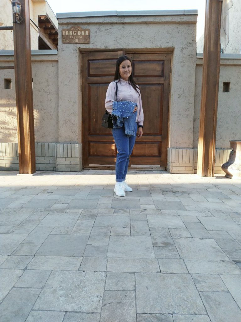 Central Asian young woman in a white collared top and blue jeans in front of a stone historic looking building with a wooden door. 