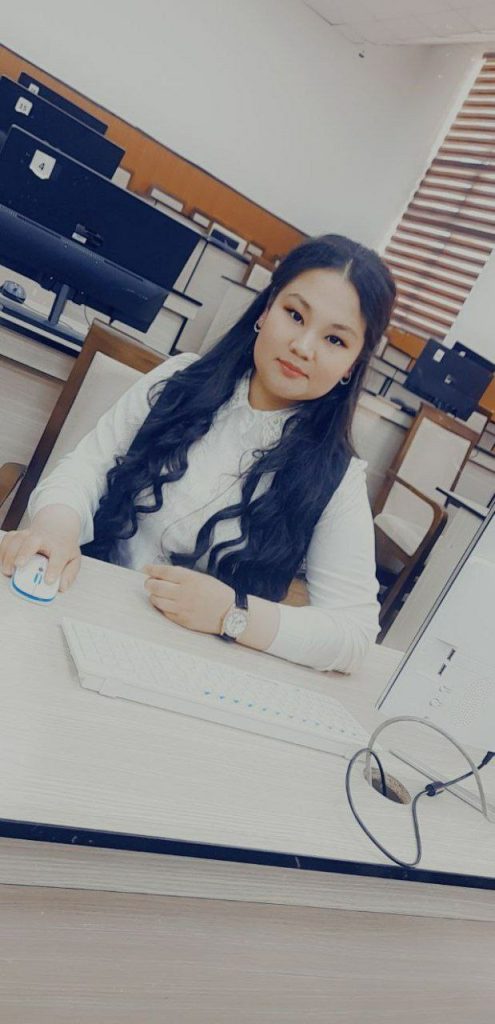 Young Central Asian woman with long dark hair in a room with computers seated at a desk. 
