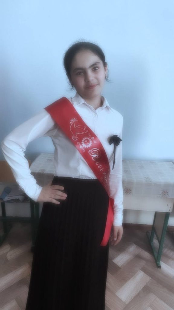 Central Asian teen girl with dark hair up in a bun and a white collared shirt and dark pants and a red sash.