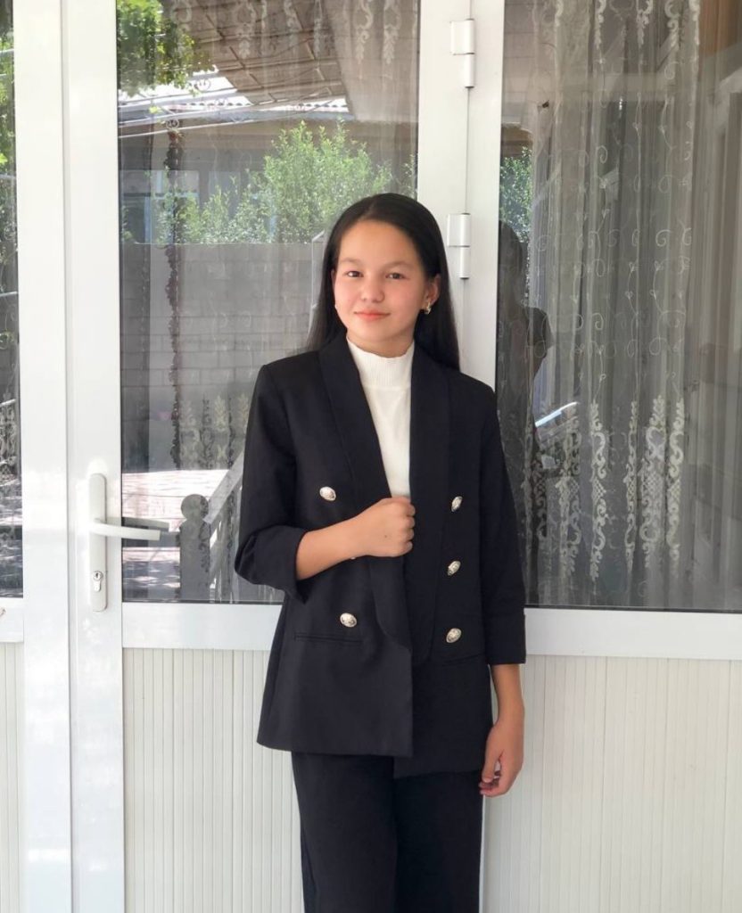 Central Asian teen girl with long dark coat, white top, and black pants, standing in front of a window.