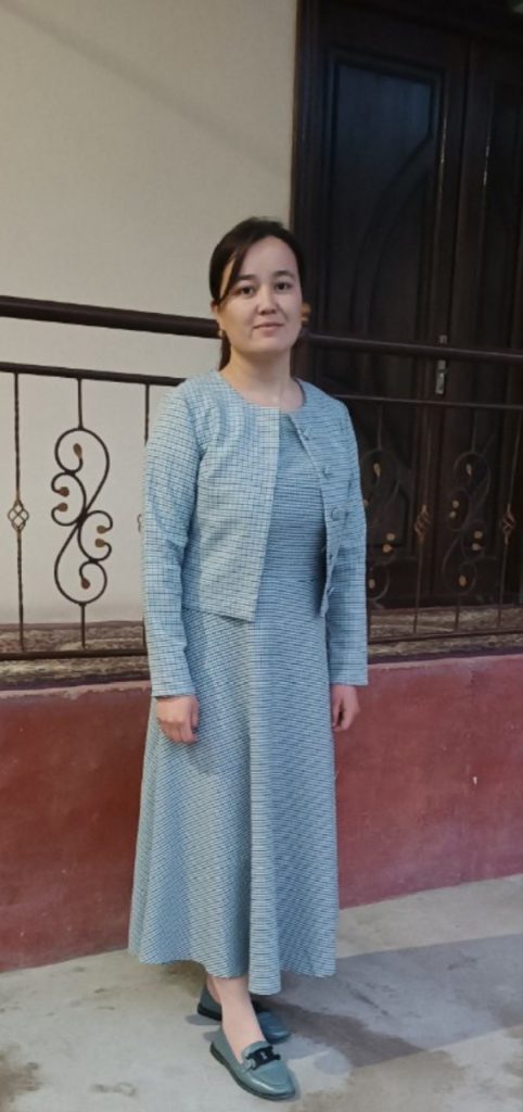 Young Central Asian woman standing outside a door in a long blue dress and coat with buttons.