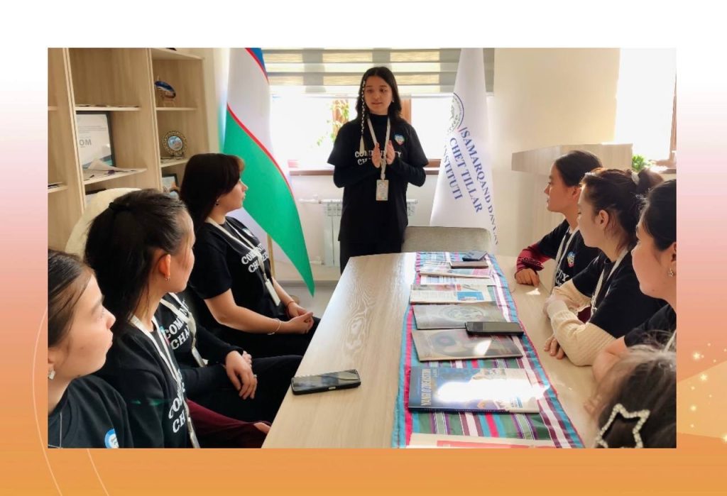 Young Central Asian teen girls seated at a classroom table listening to one of them standing to speak to them. Uzbek flags and bookshelves in the background.