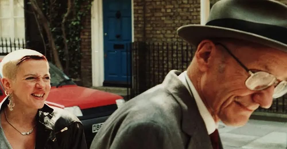 Image of Kandy Fontaine, short haired middle aged white woman, standing on a city street near a car and brick buildings and an older white man in a suit and gray hat and reading glasses.