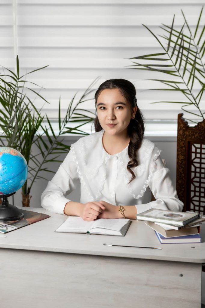 Young Central Asian woman with dark curly hair and a white ruffled blouse seated at a table with a book open in front of her. She's got a globe and a magazine and some leafy green plants behind her.