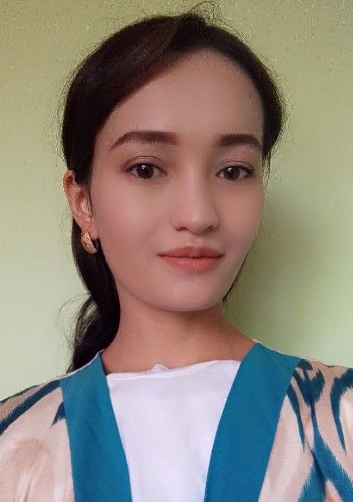Young Central Asian woman with earrings and a tan and green coat over a white blouse.