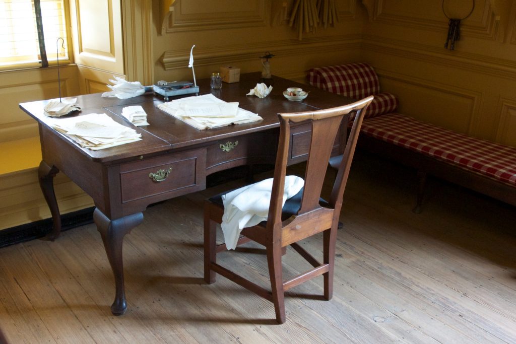 Old style writing desk with a wooden chair and wood floor and papers for an ink pen. Window and cot in the room. 