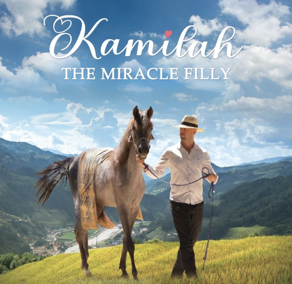 Movie poster for Kamilah the Miracle Filly. Man in a white shirt and wide brimmed hat leading a tall racehorse through a mountainous area with a vista. Sunny day with a few clouds in the sky. 