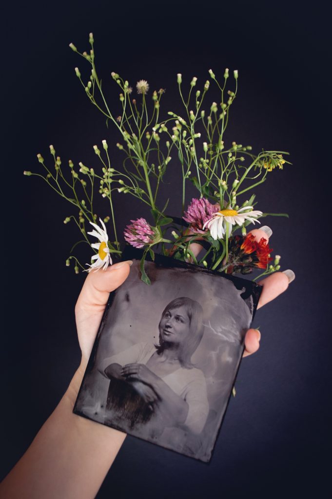 Person's hand holding an old black and white photograph and some flowers. Daisies, baby's breath, a pink flower. 