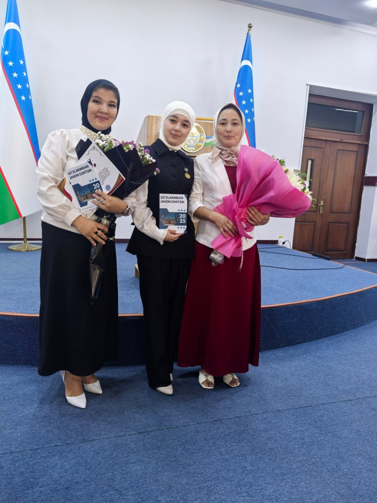 Three young Central Asian girls holding certificates and flowers standing on a blue stage near an Uzbek flag. 