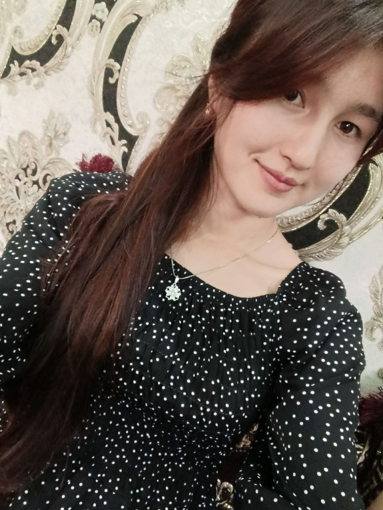 Central Asian young woman with long dark hair and a necklace and black top with white polka dots. 