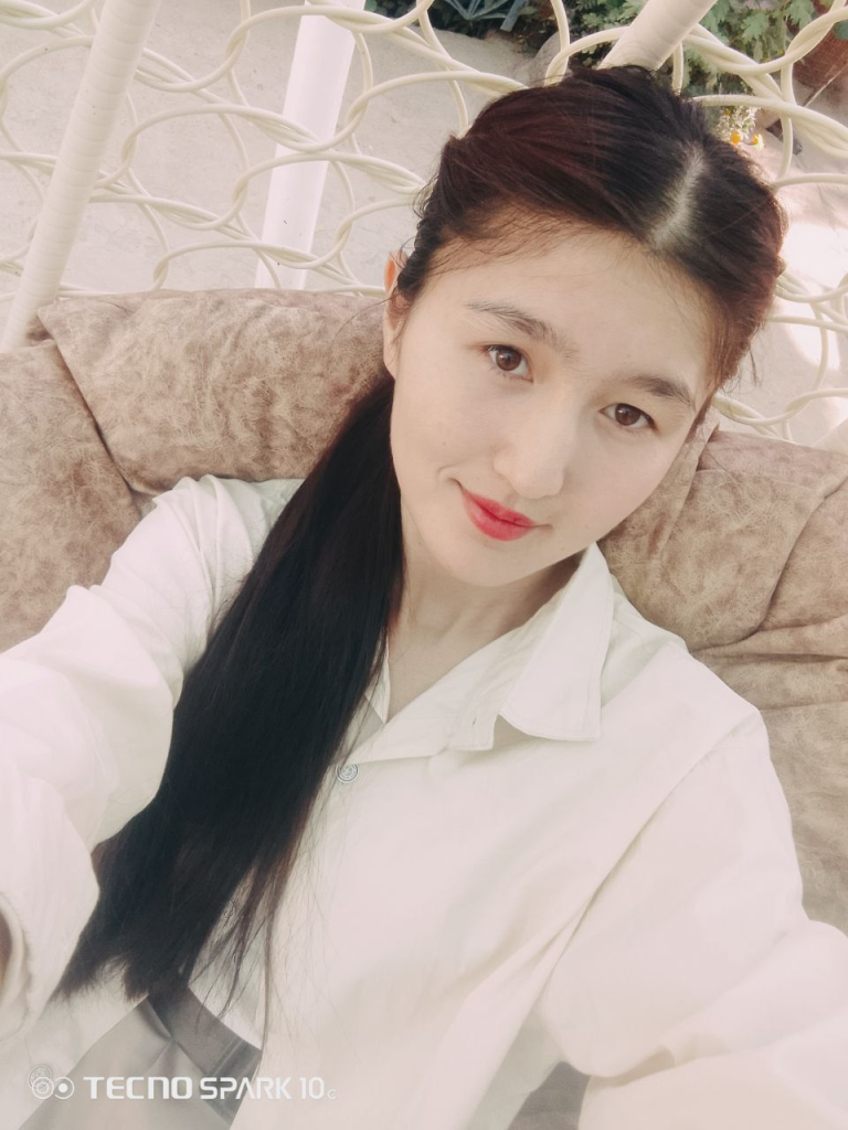 Young Central Asian woman with long dark hair, brown eyes, and a white blouse, seated on a tan patterned couch.