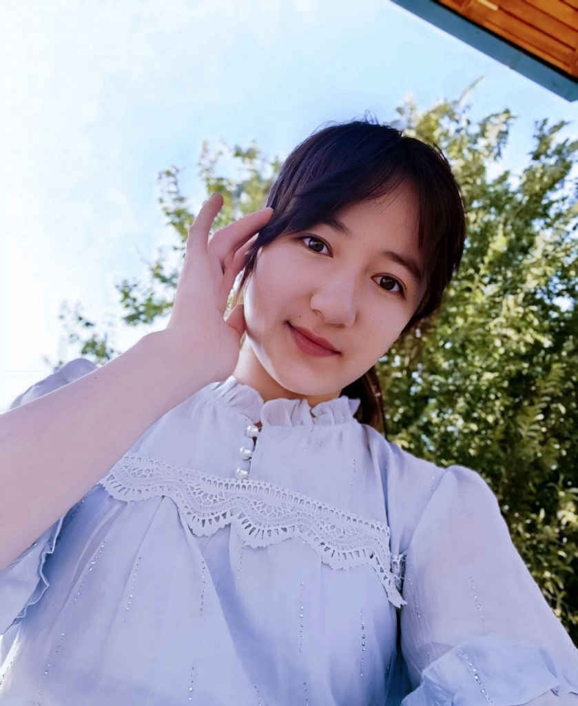 Young Central Asian woman with long dark hair, brown eyes, and a white ruffled blouse outside in front of a leafy green tree on a sunny day.
