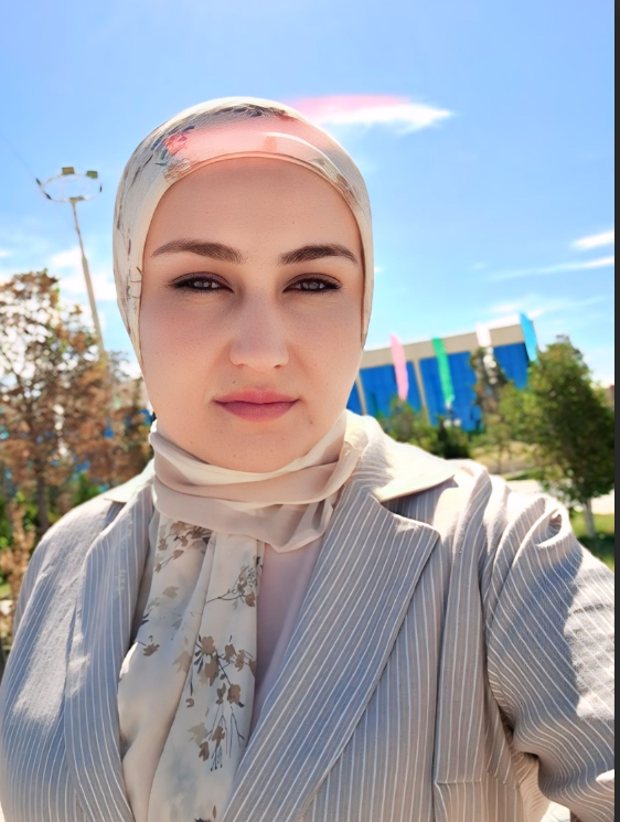 Young Central Asian woman with a light flowered headscarf, light pink top and gray and white striped coat. She's outside a building on a sunny day with trees and a few clouds in the sky.