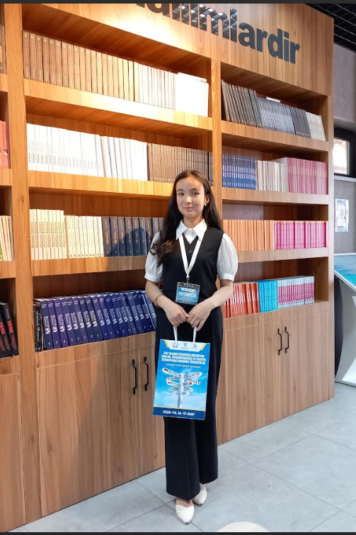 Young Central Asian woman in a black vest and white blouse with a bookshelf behind her and a lanyard around her neck.
