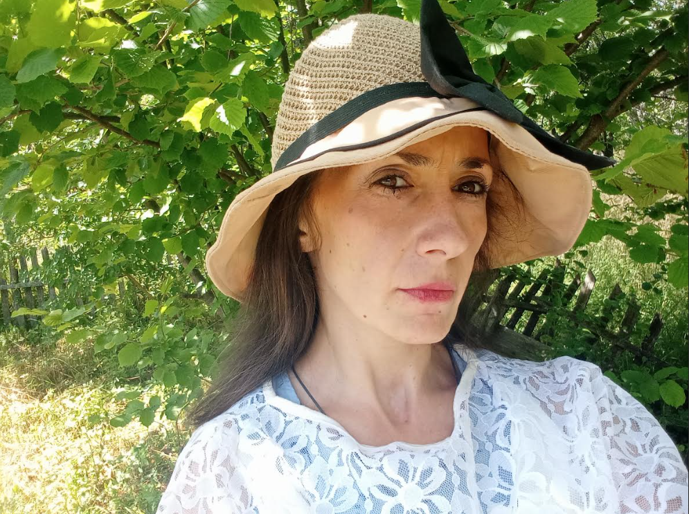 Middle aged light skinned European woman with a big straw hat and white blouse standing on a green lawn in front of a leafy green tree.