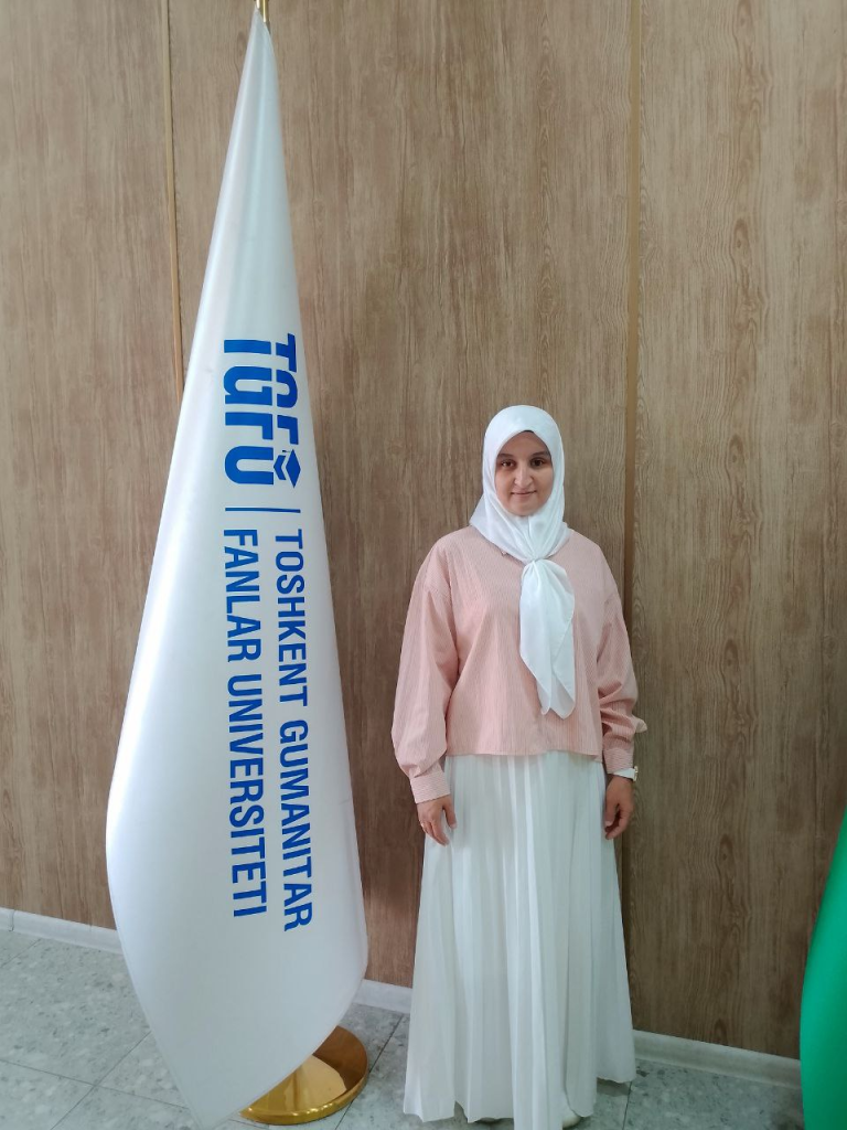 Young Central Asian woman in a white headscarf and pink sweater over a white dress. She's standing near a blue and white flag for some organization. 