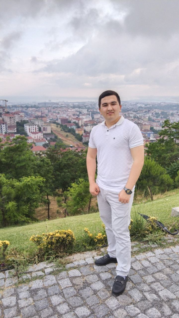 Young Central Asian man standing in front of trees and green bushes and a view of a city with tall buildings. 