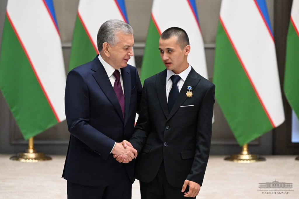 Young Central Asian man with very short hair and a black suit and pants shakes the hand on stage of a similarly dressed older man. Uzbek flags on stage. 