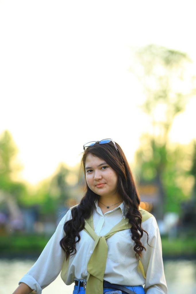 Young Central Asian woman with sunglasses on her head and a collared shirt standing outside in the grass on a sunny day. She's got a yellow scarf and jeans. 