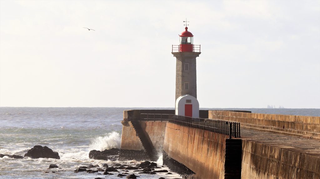 Concrete pathway to a lighthouse with a red door and the ocean and rocks in the distance.