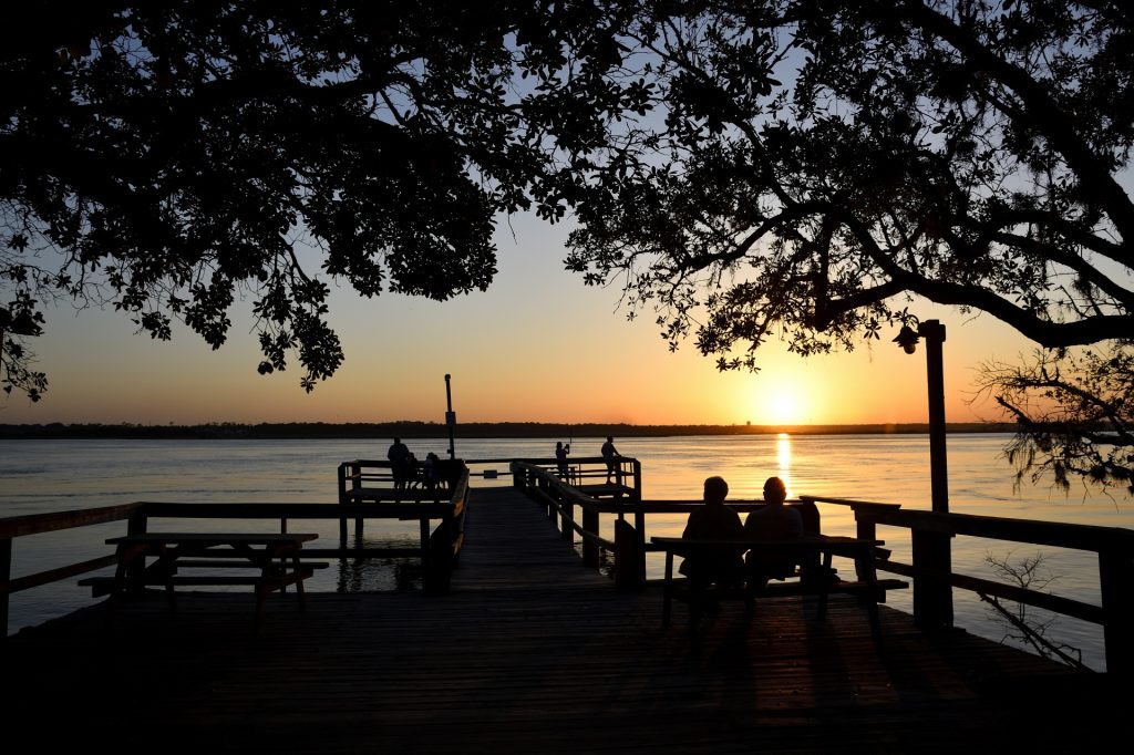 Calm water with sunset/sunrise and silhouettes of a wooden pier, trees, and two people watching.