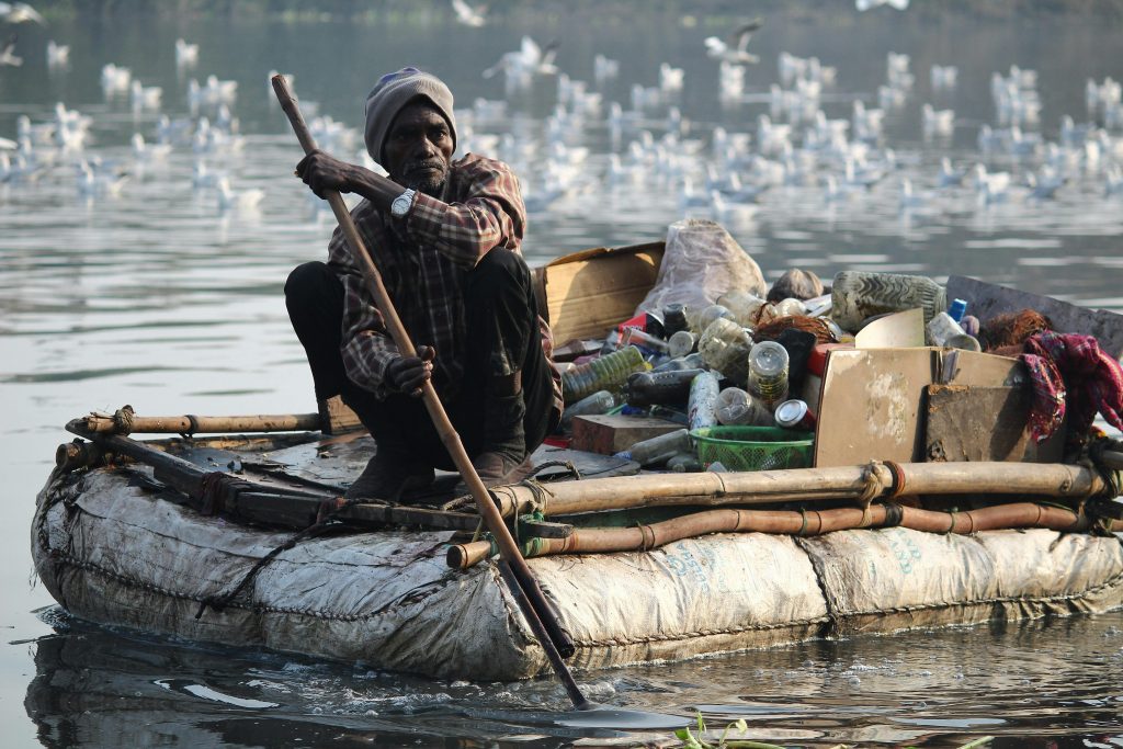 Middle aged South Asian man on a modest raft carrying boxes full of bottles pushing himself down the river with a pole. White birds in the background.