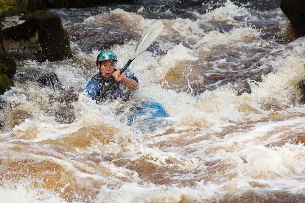 Young man on a blue kayak with a paddle navigating through rocks and white water.