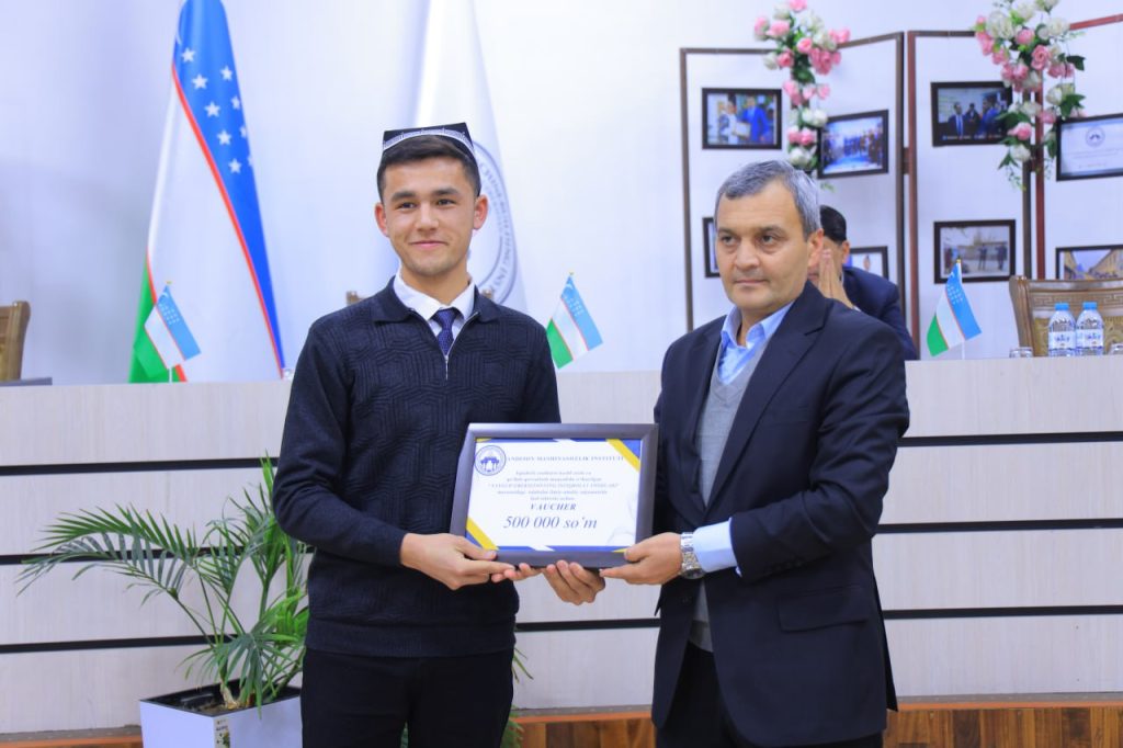 Author (left) being awarded a certificate from an older man (right). Both are Central Asian men in dark suits and ties.  Flag and green leafy plant in background. 