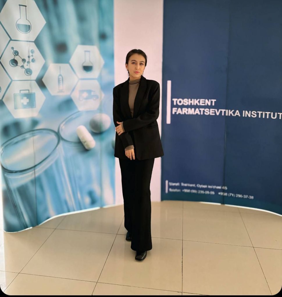 Central Asian woman standing in front of a set of medical research themed posters. She's got dark hair and a black coat and black pants over a light tan top.