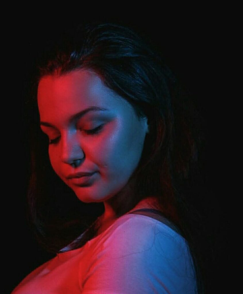 Young Eastern European woman looking to the left with her eyes closed in a dark room. She's in a light colored blouse and has short dark hair.