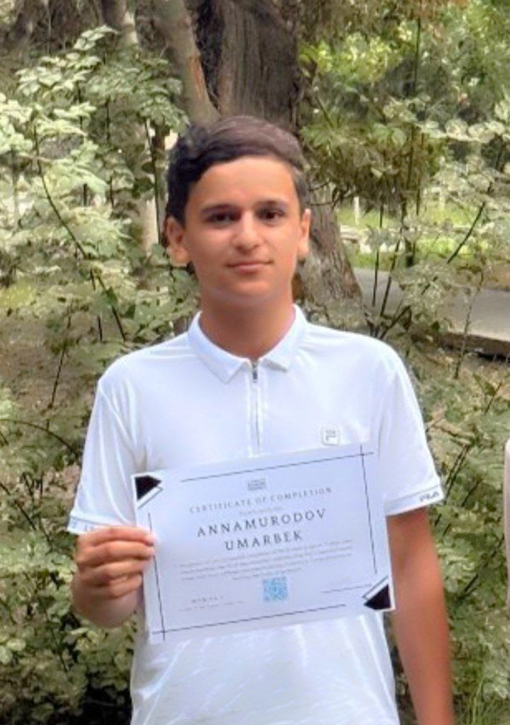 Central Asian boy with short dark hair and a white collared shirt holding a certificate and standing in front of greenery and a tree with a large trunk.