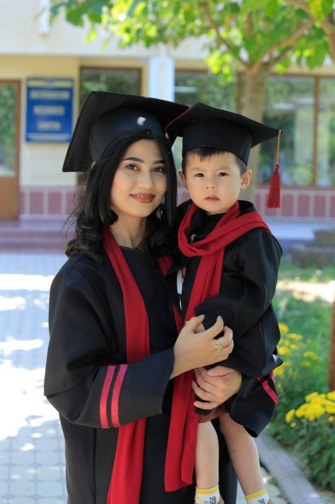 Young Central Asian woman in a black cap and gown and red sash holding a small boy in the same outfit. She's outside a building on a sunny day by a leafy tree. 