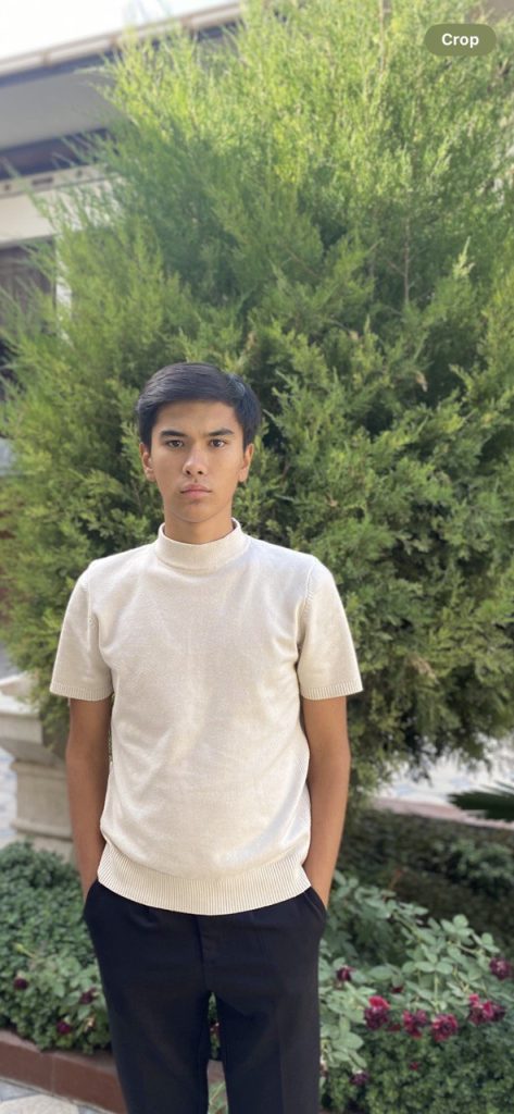 Young Central Asian man with short dark hair and a white tee shirt standing in front of a juniper bush. 