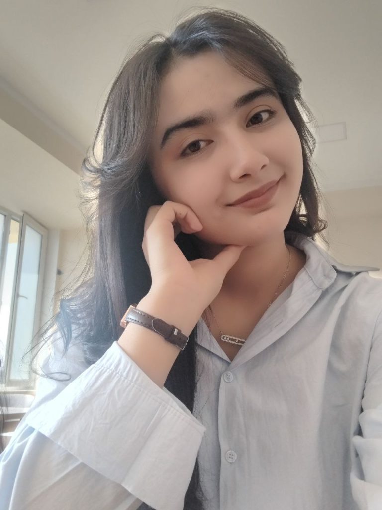 Young Central Asian woman with brown eyes and hair, resting her head on her hand and wearing a light-colored blouse and a brown watch. 